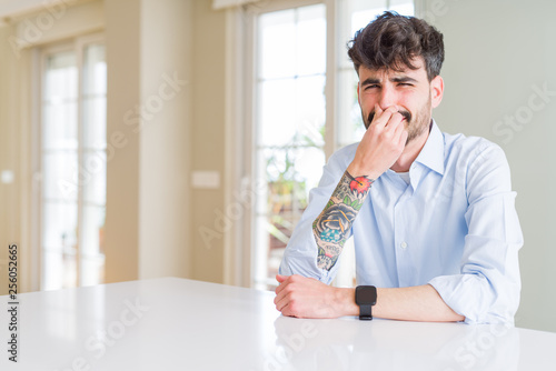 Young businesss man sitting on white table smelling something stinky and disgusting, intolerable smell, holding breath with fingers on nose. Bad smells concept.