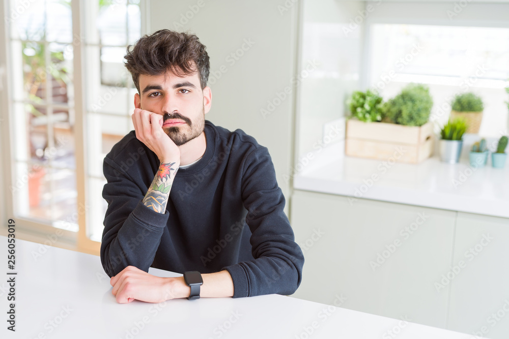 Young man wearing casual sweatshirt sitting on white table thinking looking tired and bored with depression problems with crossed arms. Young man wearing casual sweatshirt sitting on white table thinking looking tired and bored with depression problems with crossed arms.