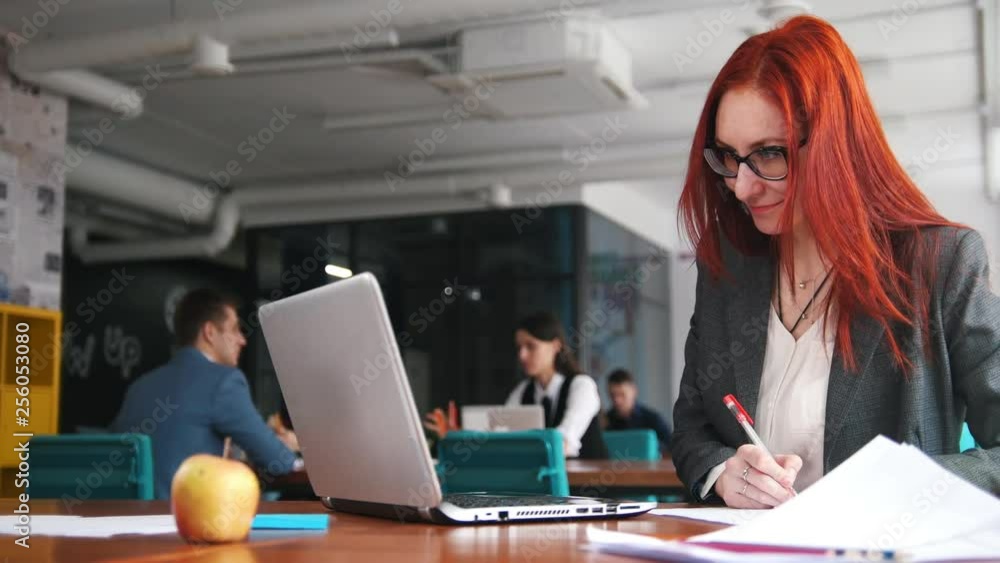 A ginger woman sitting at office and working by the laptop and making notes A ginger woman sitting at office and working by the laptop and making notes