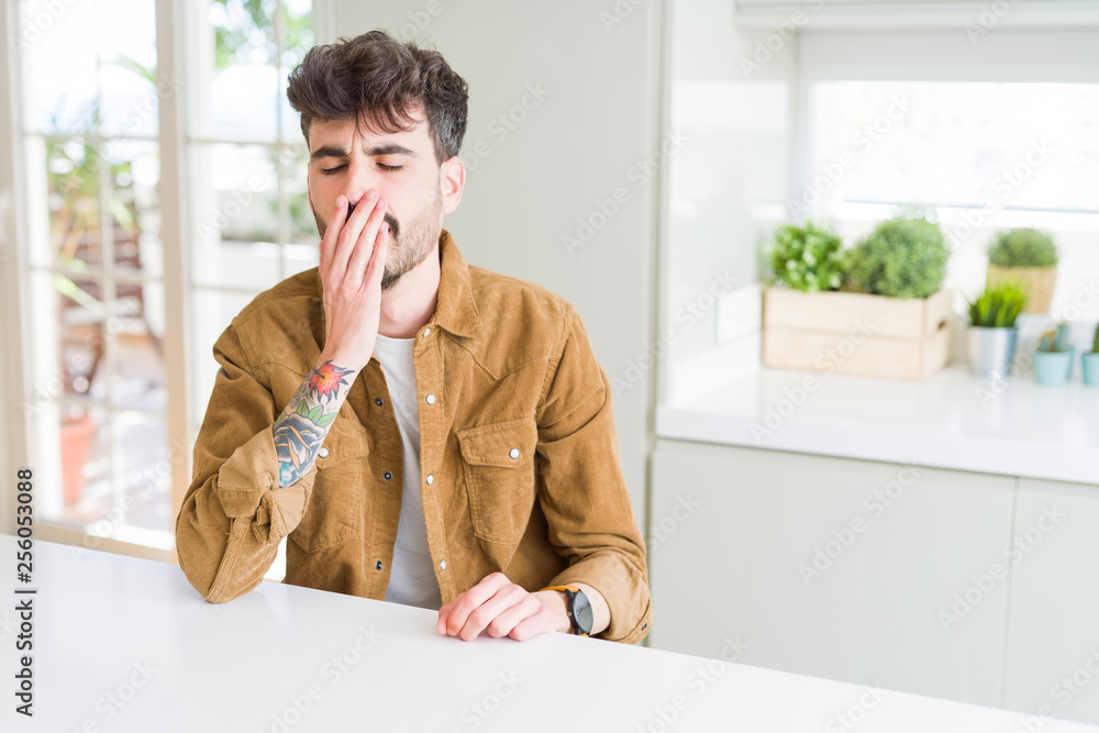 Naklejka premium Young man wearing casual jacket sitting on white table bored yawning tired covering mouth with hand. Restless and sleepiness.