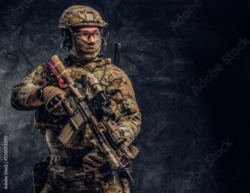 Fully equipped soldier in camouflage uniform holding an assault rifle. Studio photo against a dark textured wall