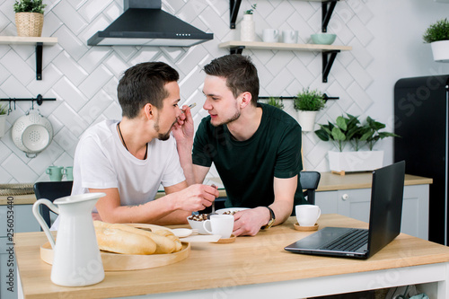 Two happy young Caucasian gay men looking at one another with smiles during breakfast. Man feeding his partner. Happy morning  gay couple concept