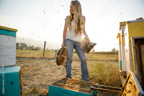 Woman beekeeper stand by hive boxes and smiles while bees fly around