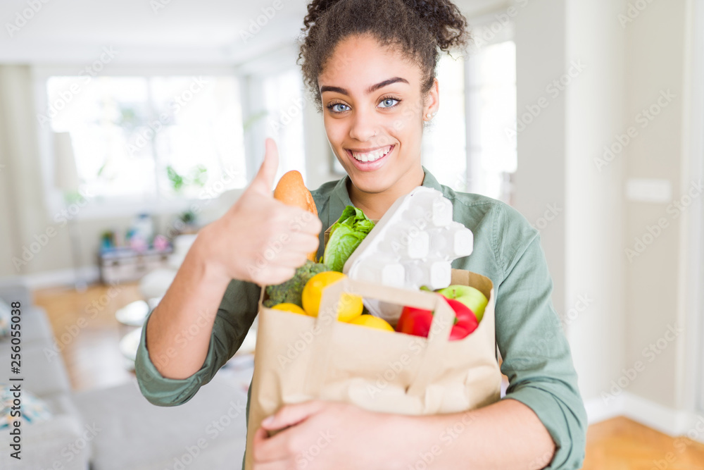 Young african american girl holding paper bag of groceries from supermarket happy with big smile doing ok sign, thumb up with fingers, excellent sign Young african american girl holding paper bag of groceries from supermarket happy with big smile doing ok sign, thumb up with fingers, excellent sign