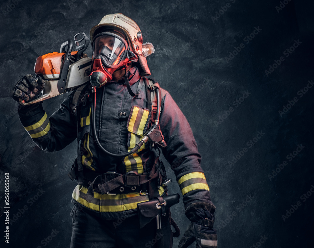 A brave firefighter wearing full protective equipment posing with a chainsaw on his shoulder. Studio photo against a dark textured wall