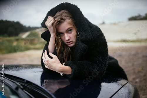 Portrait of young woman in black fur coat leaning on car bonnet photo