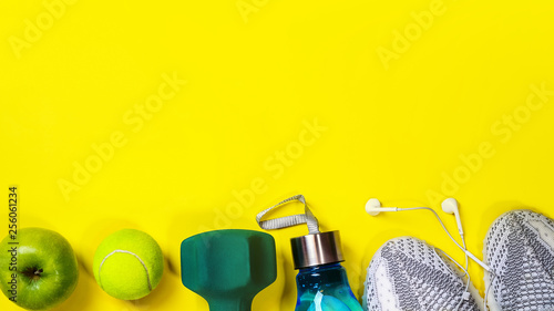 Fitness equipment and space for text on bright yellow background. Sport concept with sneakers, dumbbell, bottle of water, apple, tennis ball, headphones. Copy space, flat lay.Top view, banner.
