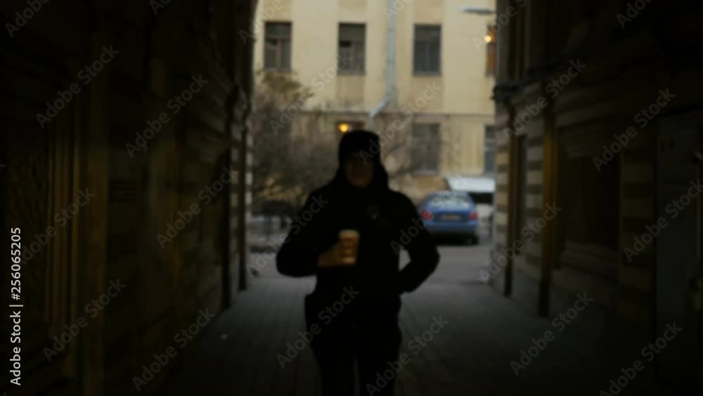 Front view of young man walking down the city street and drinking coffee. Serious tired man in black walks on the street. Front view of young man walking down the city street and drinking coffee. Serious tired man in black walks on the street.