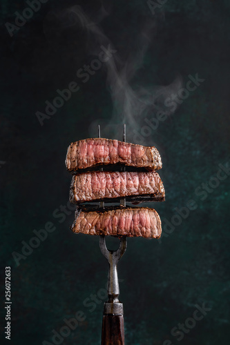 Slices of beef steak with steam on vintage fork on dark background photo