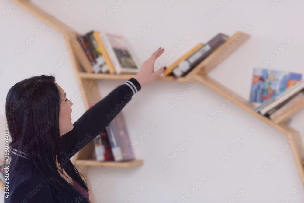 Naklejka premium Pretty young female university student choosing books from the bookshelf in the private modern library.