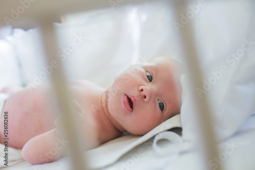 The newborn is lying in a crib with white rods. The child looks through the bars of the white bed. Meaningful look of a newborn. A healthy baby takes air baths.