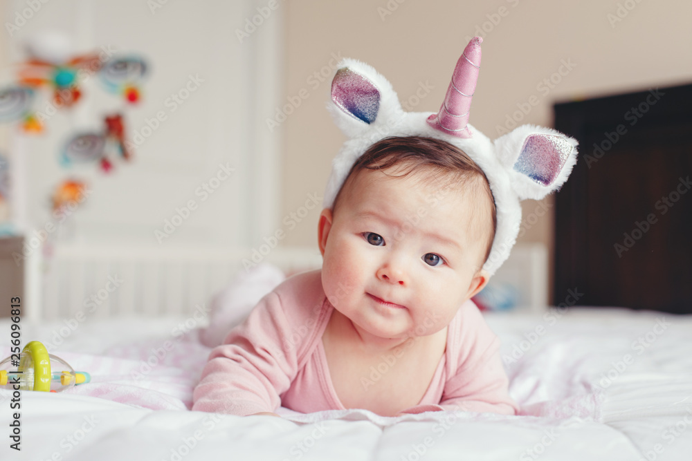 Portrait of cute adorable Asian mixed race smiling baby girl four months old lying on tummy on bed in bedroom wearing unicorn headband horn and ears looking in camera. - obrazy, fototapety, plakaty 