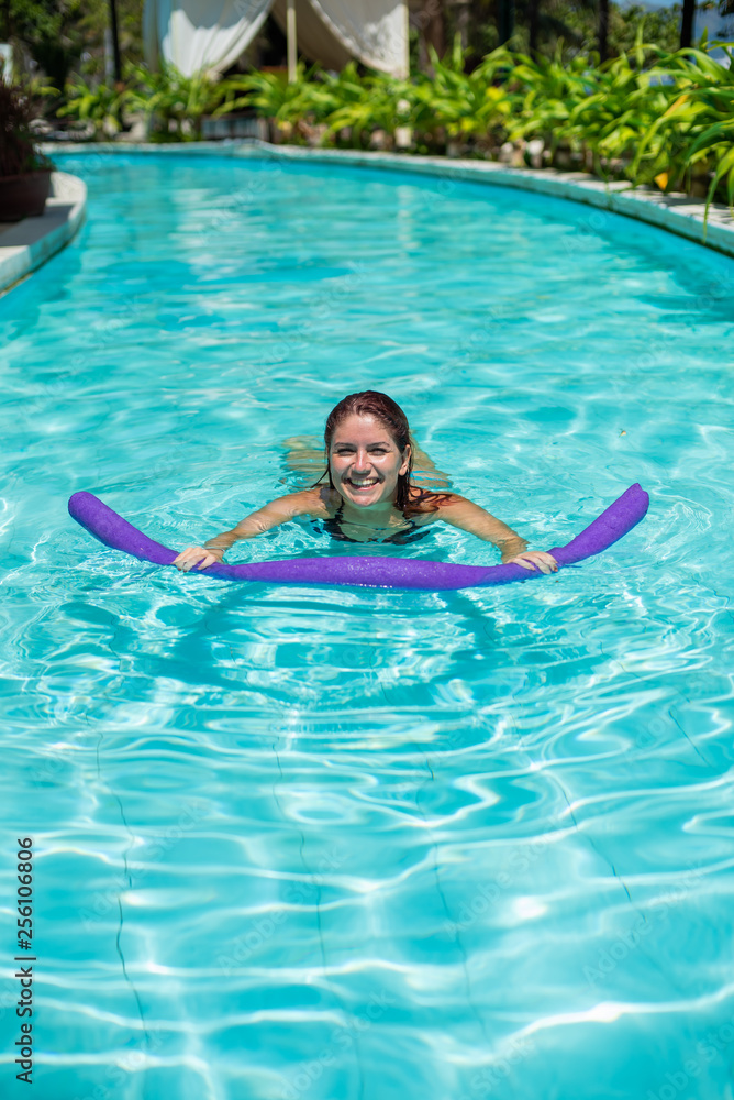 A young woman learns to swim in an outdoor pool using a swimming noodle.