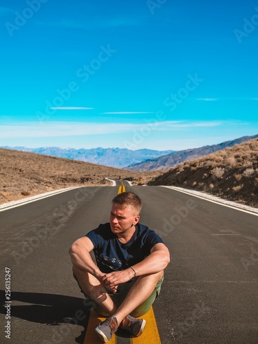 Young blond man sitting on an Empty picturesque deserted road in Death Valley  going to the beautiful mountains