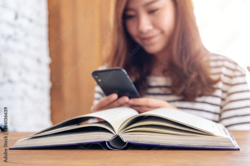 Closeup image of a beautiful asian woman holding , using and looking at smart phone with a book on wooden table Closeup image of a beautiful asian woman holding , using and looking at smart phone with a book on wooden table
