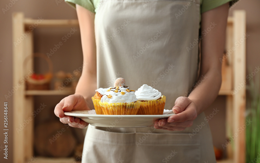 Woman holding plate with tasty Easter cupcakes in kitchen