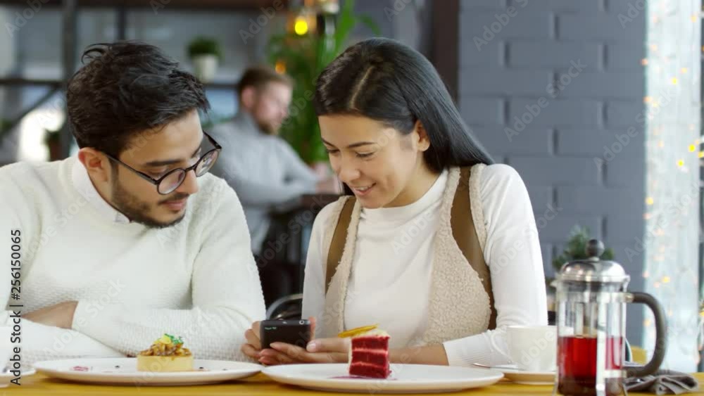 Tracking shot of young cheerful Asian woman showing something on smartphone screen to middle eastern boyfriend, smiling and talking while sitting at cafe table with desserts and tea cups on Tracking shot of young cheerful Asian woman showing something on smartphone screen to middle eastern boyfriend, smiling and talking while sitting at cafe table with desserts and tea cups on