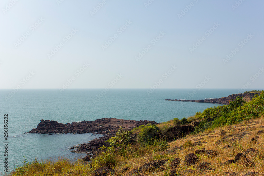 hillside with dry and green grass against a blue ocean with a rocky shore in the sea