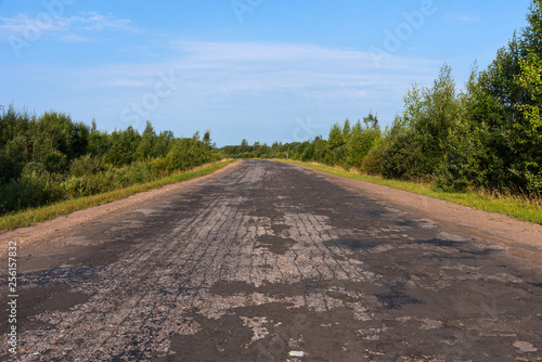 Broken empty asphalt country road surrounded by trees in sunny summer day.