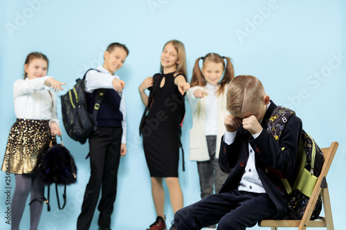 Little boy sitting alone on chair and suffering an act of bullying while children mocking. Sad young schoolboy sitting on studio against blue background.