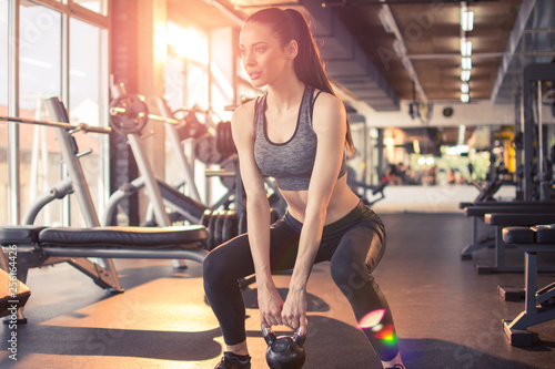 Attractive young sportswoman doing squats with kettlebell in gym