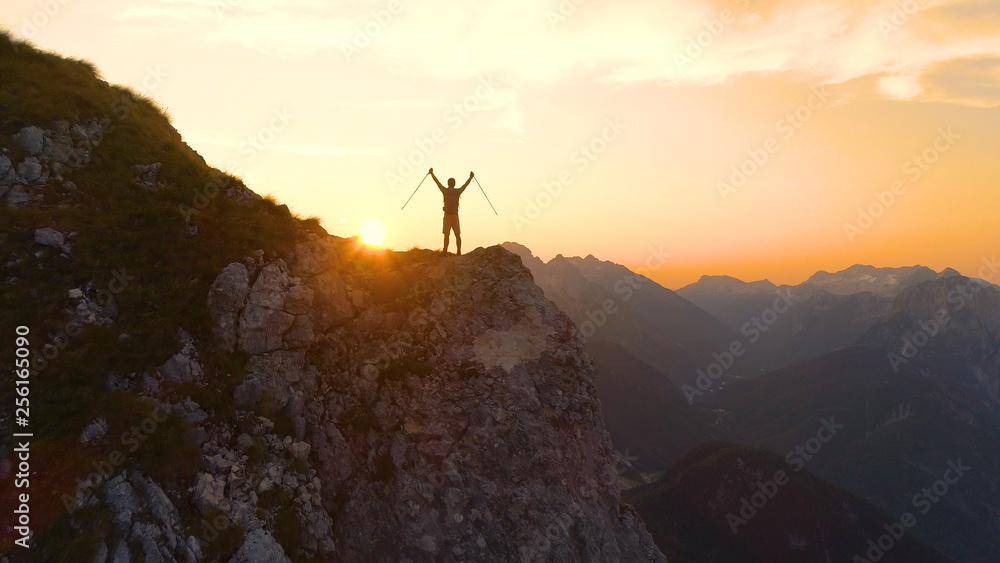 Obraz premium AERIAL: Flying behind man celebrating a successful ascent onto a scenic cliff.