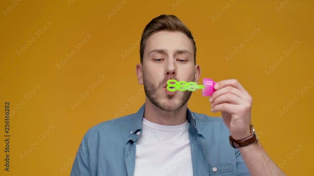 handsome man blowing soap bubbles, smiling and looking at camera in slow motion isolated on yellow handsome man blowing soap bubbles, smiling and looking at camera in slow motion isolated on yellow