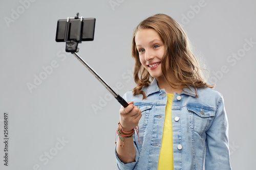 technology and people concept - happy smiling teenage girl taking picture by smartphone on selfie stick over grey background