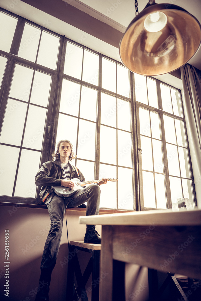 Slim rock musician wearing jeans sitting near window with guitar Slim rock musician wearing jeans sitting near window with guitar