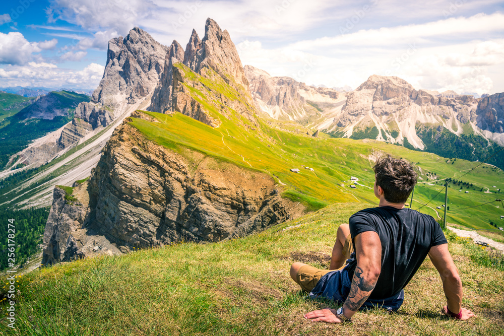 beautiful young muscular man sitting on the grass enjoying the beautiful dolomites mountains landscape. seceda, 2500m over the odle group mountains - obrazy, fototapety, plakaty 