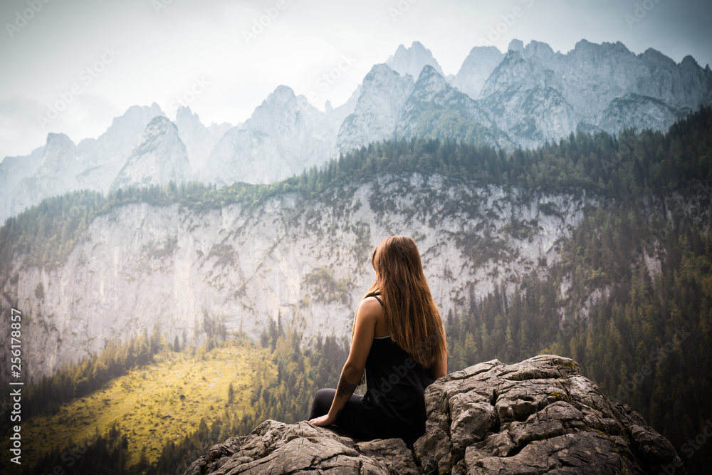 young girl with long hair sitting on the top of a rock enjoying the beautiful view of  the mountains covered by forest