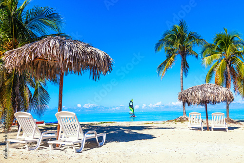 Sun loungers under straw umbrellas on the sandy beach with palms and sailboats near ocean and sky. Vacation background. Idyllic beach landscape.