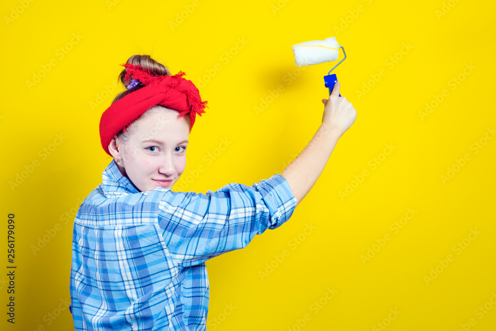 Young beautiful girl in a plaid shirt and a red bandage on his head, paints with a paint roller against the background of a yellow studio. Retro style. Young beautiful girl in a plaid shirt and a red bandage on his head, paints with a paint roller against the background of a yellow studio. Retro style.