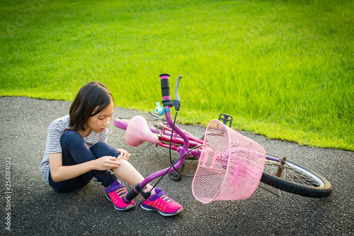 Asian little girl sitting down on the road with a leg pain due to a bicycle accident,the bike fall near the child,girl riding a bicycle with a slight wound in the park,falling bicycle,Acciden concept photo