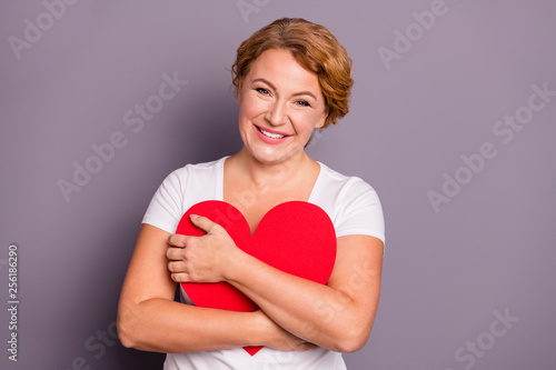 Portrait of nice cute lovely attractive tender cheerful cheery wavy-haired lady in casual white t-shirt holding in hands cuddling sweet heart isolated on gray violet purple pastel background