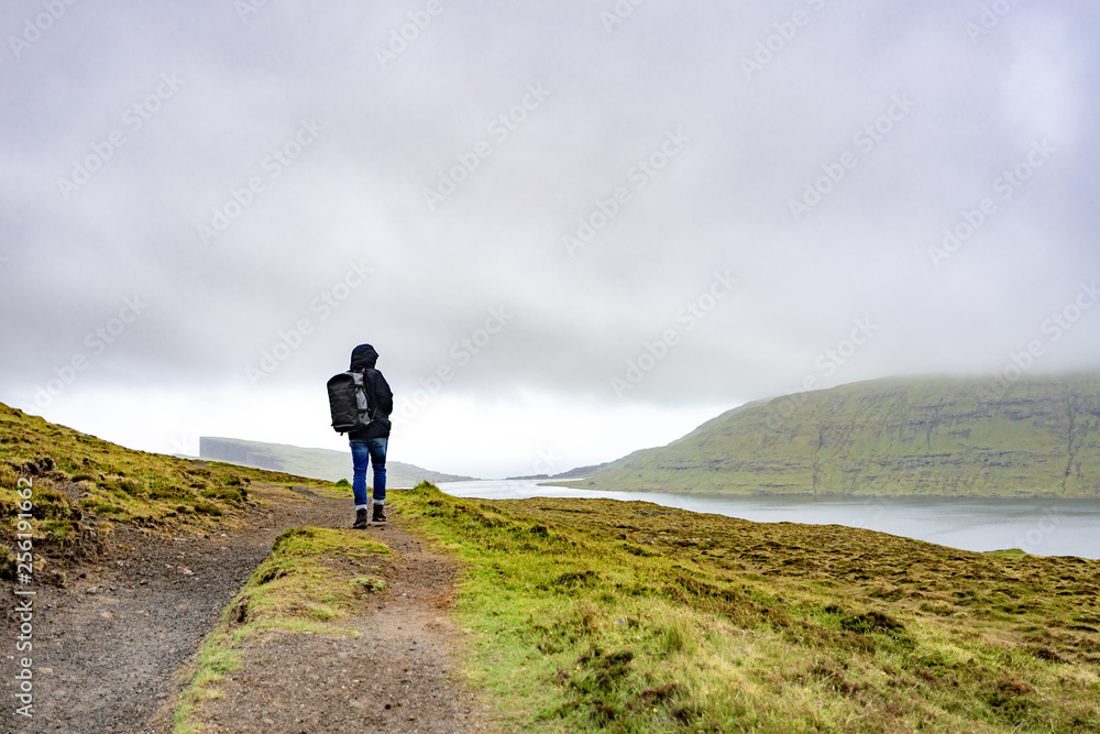 Young man walking or trekking on rural road beside the lake with bad weather raining in Faroe Islands, north Atlantic ocean, Europe