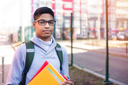 Portrait of African American student in glasses with books. Black young man smiling is standing on the street holding textbooks in his hands. 