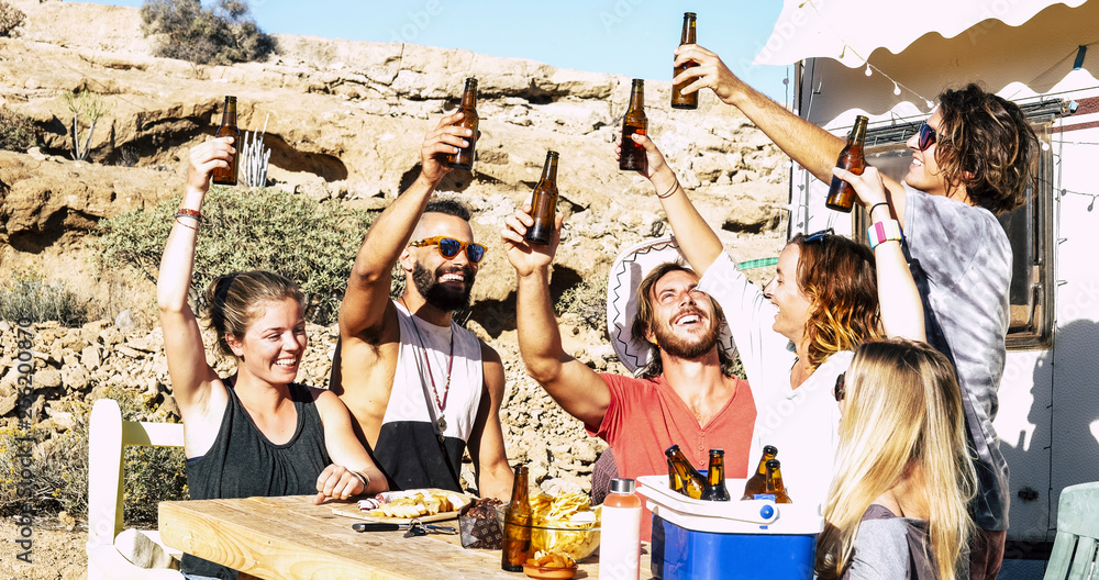 Fun success and friendship concept with group of young men and women people all together toasting with beers and food sitting on a rural wood table with nature in background
