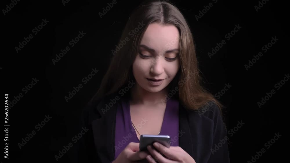 Closeup shoot of young attractive caucasian female texting on the phone looking at camera and smiling playfully with background isolated on black Closeup shoot of young attractive caucasian female texting on the phone looking at camera and smiling playfully with background isolated on black