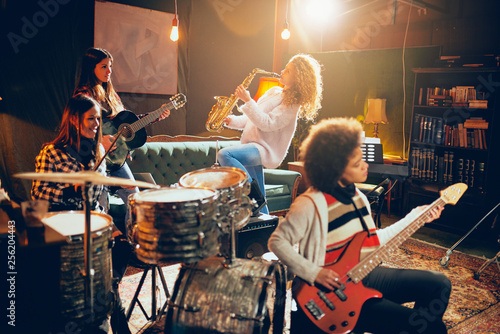 Girls playing jazz music. In foreground one woman playing bass guitar and in background other two playing saxophone and drums. Home studio interior.