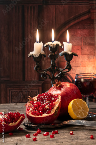 Still life in a rustic style. Fruit-pomegranates, lemon lying on a wooden table with a glass of wine and candles on the fireplace background