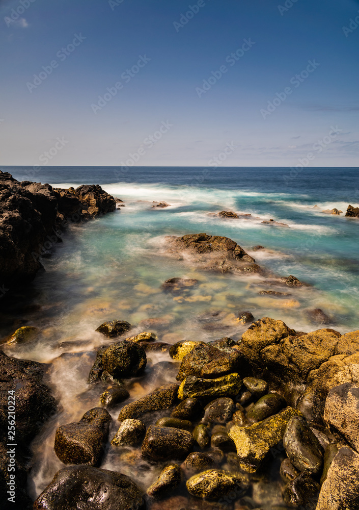 Obraz premium El Sauzal volcanic coastline, long exposure photography, with afternoon sunlight, blue ocean, horizon and blue sky, with few small clouds, Tenerife, Canary islands, Spain