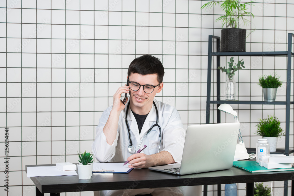 Male doctor using telephone while working on computer at table in clinic Male doctor using telephone while working on computer at table in clinic