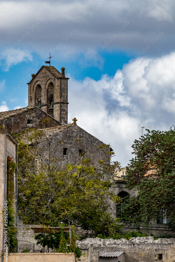 Church tower with two brass bells, stone side wall and roof with religious cross, street view of ancient town of Matera, Basilicata, Southern Italy, cloudy summer warm August day