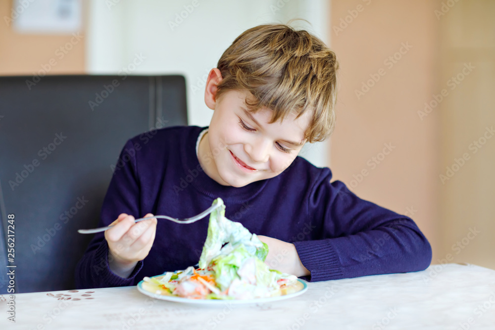 Happy kid boy eating fresh salad with tomato, cucumber and different vegetables as meal or snack. Healthy child enjoying tasty and fresh food at home or at school canteen.