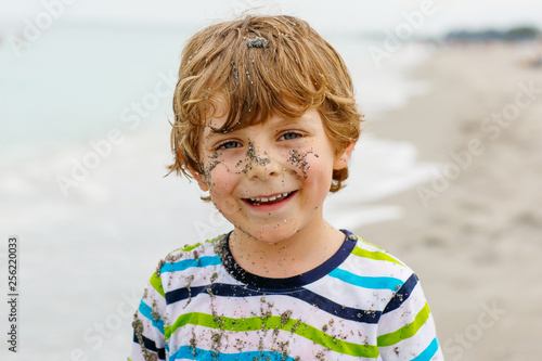 Adorable active little kid boy having fun on beach of North Sea in Germany. Happy cute child relaxing, playing and enjoying stormy warm day near palms and ocean. Kid with sand on funny beautiful face.