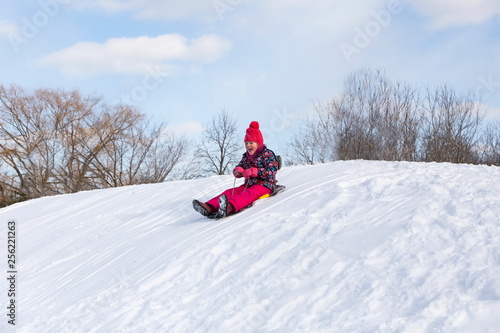 Pretty little girl in her ski suit screaming of joy while starting to slide down a small snow covered hill with her sledge board during a bright sunny winter day
