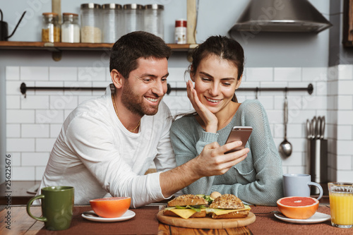 Happy young loving couple at the kitchen have a breakfast using mobile phone.
