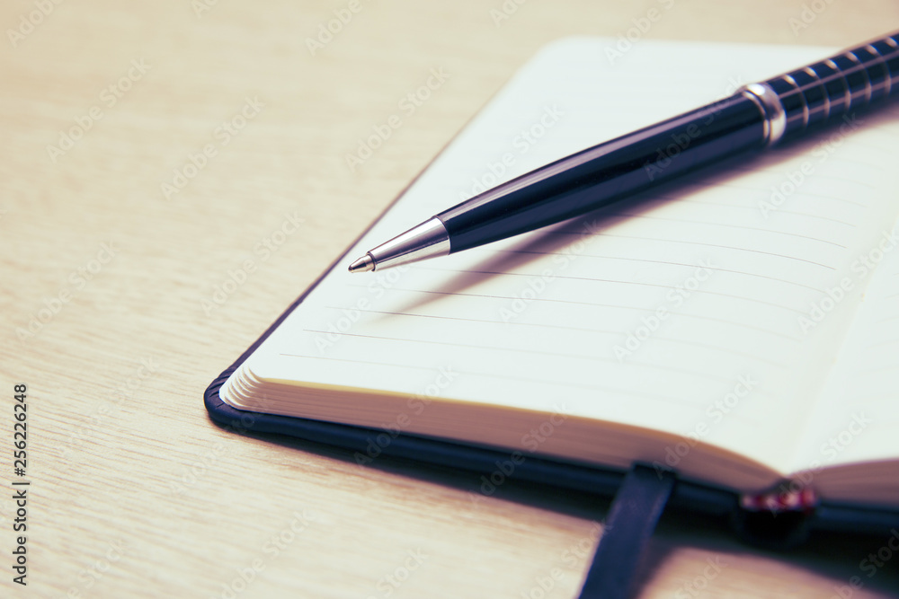 Black pen lies on the opened note cover on the spring on a light wooden table. Place to write. Black pen lies on the opened note cover on the spring on a light wooden table. Place to write.
