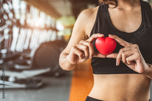 Fototapeta Naklejka Na Ścianę i Meble -  Happy sport woman holding red heart in fitness gym club. Medical cardio heart strength training lifestyle. Pretty female sport girl workout exercise. Cardiac healthy and well-being. Massage ball in ha